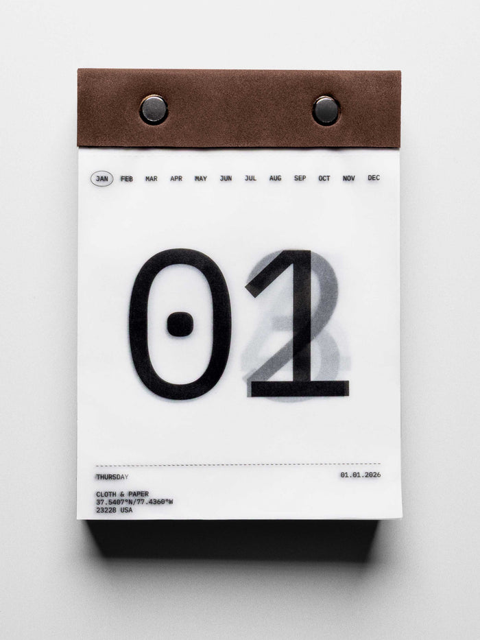 Vellum paper calendar tear-off desk pad bound with suede and metal pins. Page shows January 1st in large typographic design with days below slightly visible through transparent paper.
