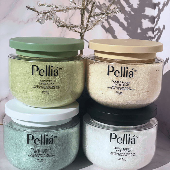 Four jars of Pellia bath soaks stacked on a marble surface with a decorative plant in the background.